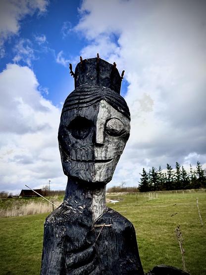 The image features a wooden sculpture of a figure with a striking and somewhat stylized design. The sculpture has a large, elongated head with detailed facial features including eyes, a nose, and a mouth etched into the wood. The head is adorned with multiple protruding elements that resemble spikes or nails, creating a crown-like effect. The body of the sculpture is slender and elongated, with arms flat against its sides, also carved with details that suggest clothing or armor.

The background shows a natural, outdoor setting with a clear sky and some clouds, along with grass and distant trees, indicating that the sculpture is placed in an open landscape. The art style seems rustic and possibly inspired by tribal or ancient artwork, giving it a powerful and evocative presence.