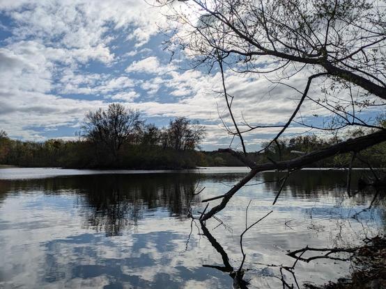 Photo of a lake with a small island near the far shore. The sky is blue and mottled by white clouds. Everything reflected on the surface of the lake.