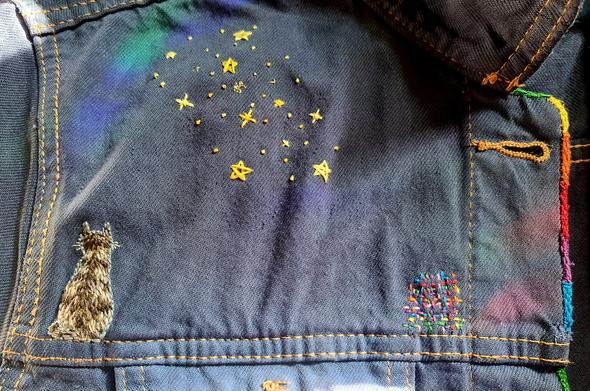 Zu sehen ist die dunkelblaue Schulterpartie einer Jeansjacke.
Der obere Teil ist mit einem Sternenhimmel in verschiedenen Gelbtönen bestickt. Auf der linken unteren Seite sitzt eine gestickte, grau getigerte Katze mit dem Rücken zum Betrachter. Auf der rechten unteren Seite ist eine kunterbunte Flickstelle in allen Regenbogenfarben. 
Auch die mit Regenbogenfarben gesäumte Knopfleiste ist rechts im Bild zu sehen.
Ein Sonnenfänger wirft einige  Regenbogenlichter auf die Jacke.