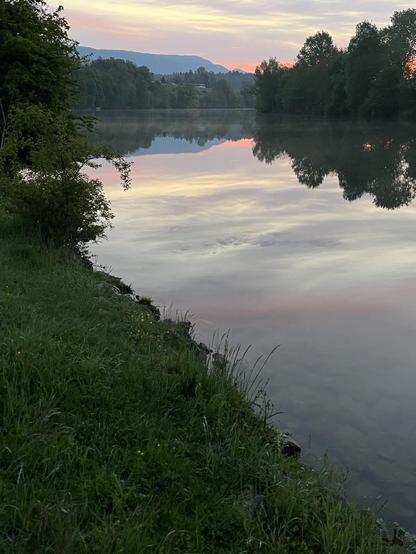 Morgenstimmung am Fluss, die Sonne geht gerade auf, der leicht wolkige Himmel spiegelt sich im ruhigen Wasser.