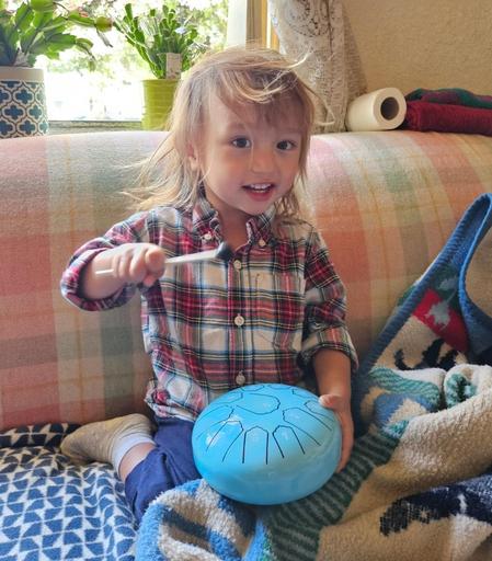 A young child with blonde hair sits cross-legged on a couch covered with blankets, holding a blue steel tongue drum. The child wears a red and white plaid shirt and navy pants, and they are holding a mallet, poised to strike the drum. They are looking at the camera with a slight smile. Sunlight streams in from the window behind them, next to plants.