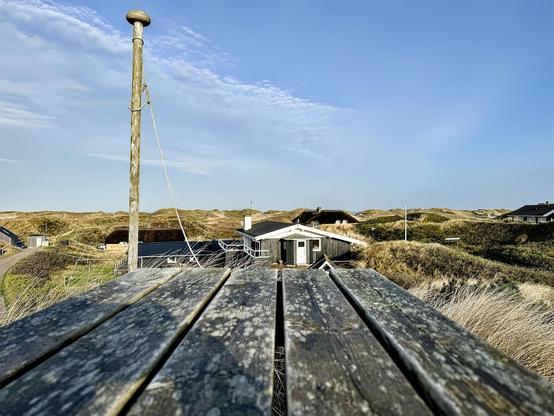 This image shows a tranquil coastal scene characterized by dune landscape. There is a weathered wooden table in the foreground, leading the eye towards a small building possibly used for housing or service facilities. The environment includes windswept grass and sand dunes under a clear blue sky. There's a rustic pole with ropes, possibly part of a basic utility or flag structure. Overall, this depicts a serene and somewhat remote setting, likely near a beach.