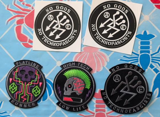 Des patchs et deux stickers. 
On y voit écrit no gods no techno fasciste. Et on voit bien les trois flèches qui pointent en bas à gauche. Mais elles ont un petit côté logo USB. 
Et deux patchs qui font penser à net runner ou au cyberpunk avec écrit high-tech low life et un autre flatline runner.
