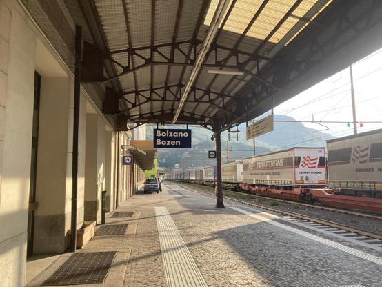 Bahnsteig 1 am Bahnhof Bolzano/Bozen, von rechts strahlt Morgenlicht ins Bild, die Frachtwaggons auf Gleis 2 werfen lange Schatten