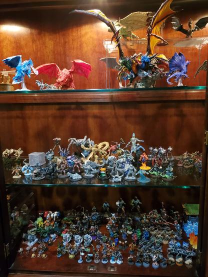 A photo of a cabinet holding 3 shelves of minis. The bottom shelf has over a hundred figures around an inch tall. They are a variety of species and wield a variety of weapons. The middle shelf has slightly larger minis. Many of them are beasts or monsters or giants or elementals. The top shelf holds about a half dozen dragons of varying sizes and colors