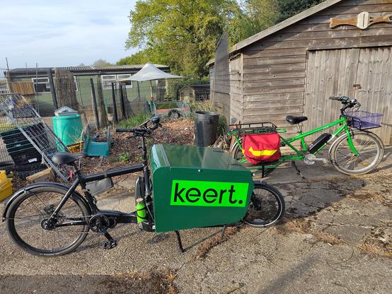 A front loading cargo bike with black frame and large green box with the Keert logo, next to a long tail cargo bike with green frame, red panniers and a purple basket, parked on broken concrete outside a wooden workshop building.
