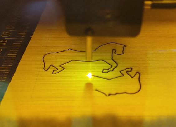 Picture of a lasercutter at work to produce tiny wooden horses. The orange protective cover causes the cutting point to glow orange