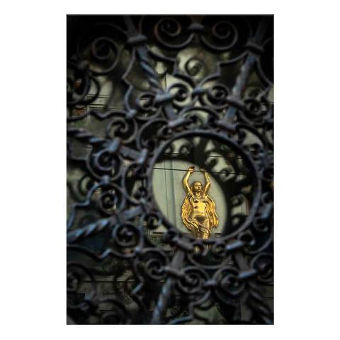 A small, barred viewing window in the doorway of a downtown house reflects the golden statue of a woman on the wall of the house opposite.