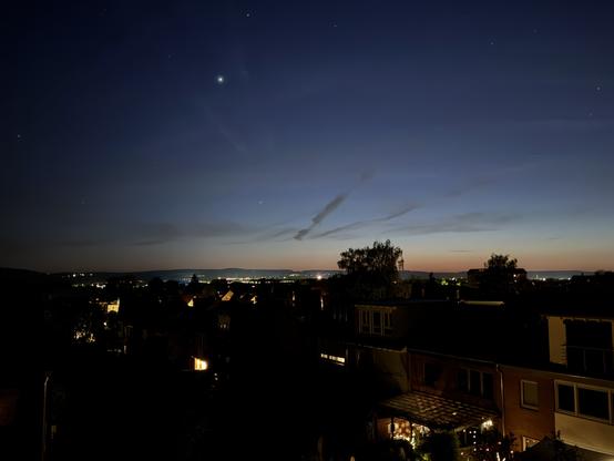 Ein nächtliches Stadtbild mit silhouettenförmigen Dächern und beleuchteten Fenstern darunter, mit einem Verlaufshimmel, der von dunkelblau zu helleren Tönen übergeht. Mehrere Sterne sind sichtbar, und ein Planet leuchtet hell im oberen Teil des Bildes.