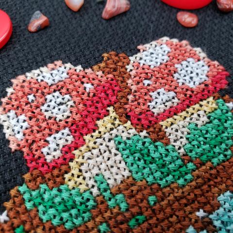 An extreme close up of a cross stitch of a two red and white mushrooms in the dirt and leaves.