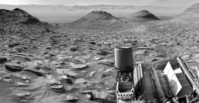 A black and white image capturing a Martian landscape, with the rear section of the Curiosity rover in the foreground. The two buttes at center are Gould Mesa and Wilkerson Butte, with the wall of the rim of Gale Crater in the background, at 10 kilometers distance.