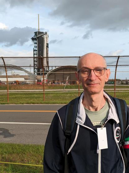 Daylight photo of a bald man with eyeglasses, wearing a green t-shirt and a navy blue sweatshirt, and standing outdoor in front of a metallic mesh fence. Behind the fence is a launch pad with a white rocket and a black service tower next to it.