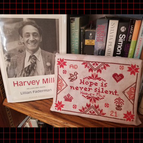 A cross stitch project and a book are staged together on a bookshelf.  Harvey Milk. His Lives and Deaths by Lillian Faderman is propped next to a quaker sampler that reads Hope is Never Silent. Harvey Milk.