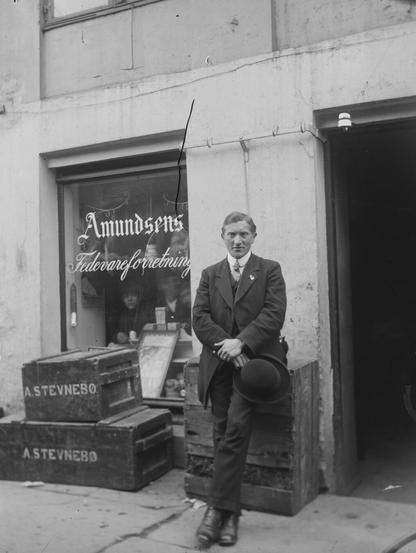 Black and white photo of man sitting outside a storefront, window reading "Amundsens fedevareforretning".