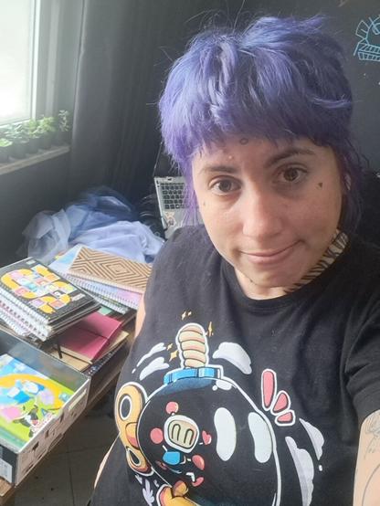 a person with purple hair color in a bomberman black t-shirt stands beside a bunch of old notebooks, two of them have adventure time on the cover