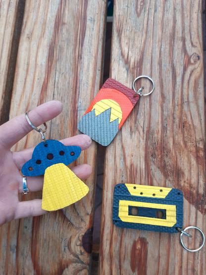 Porte-clés en lances à incendie et chambre à air de tracteur upcyclées :
- un OVNI bleu canard avec faisceau jaune
- un paysage avec montagnes kakies, soleil jaune et ciel orange et rouge
- une cassette audio bleue foncé et jaune