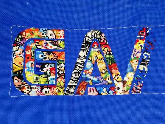 In progress cross stitch project on extremely royal blue fabric. It's spelling the word "gamer" in capitals, except only the G, A and a chunk of the first leg of the M are complete. Each letter is a collage of game characters through the ages, although without backstitching it currently just looks like a random jumble of colours in parts.