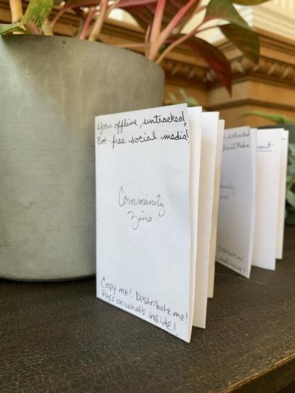 Small, handmade "zines" sit on a cafe table. Front of the paper folded accordian style says "Your offline, untracked, Bot-free social media! 
Community Zine Copy me! 
Distribute me! 
Pass on what's inside!"