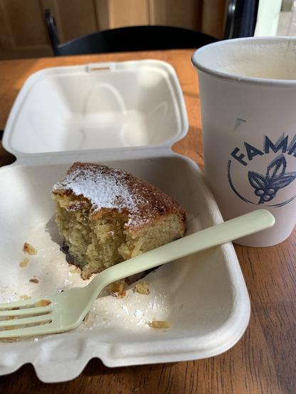 A wooden cafe table surface with a cardboard takeout container holding a half eaten piece of cake flanked by a white paper coffee cup.