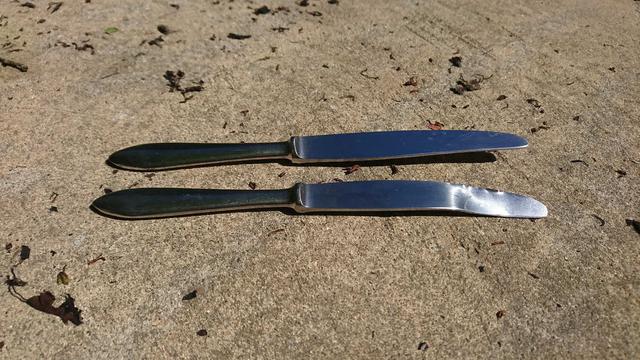 Two table knives, side by side, the tip of one is badly disformed.