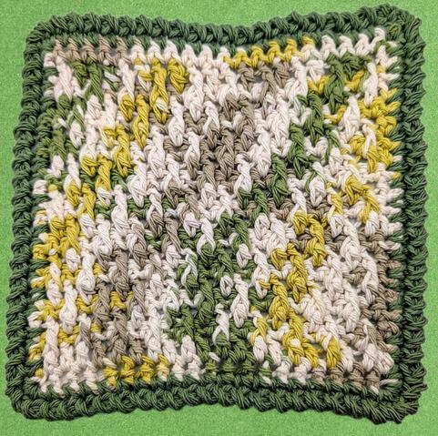 A crocheted 100% cotton dishcloth made with alpine stitch and an imitation crab stitch border. The main part is multicolored with cream, tan, yellow green, and army green, and the border is solid army green.