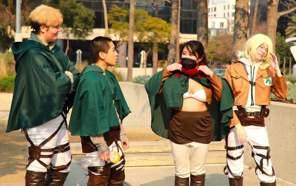 A group of four cosplayers dressed in Attack On Titan gear.  They are wearing the traditional green cloak, knee high brown boots, white pants and shirt.  One is wearing only a white bra and showing significant cleavage.