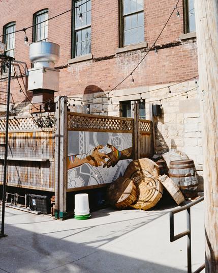 A cluttered alley in Stillwater, Minnesota, with barrels, wooden planks, and old brick walls. At the center, a colorful graffiti of two hands reaching toward each other, one painted in rainbow colors. The scene mixes industrial decay with a subtle symbol of connection and LGBTQ+ pride. Shot on 35mm film.