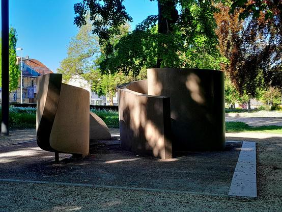 A two piece fountain. Onne side resembles an abstract snake, the other is spiral, like a snail. Skies are clear and blue, and the trees green.