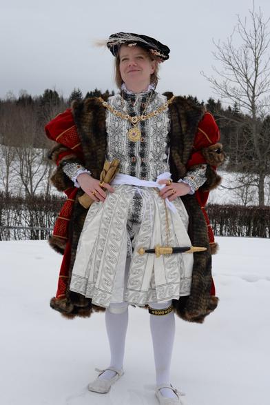 A person dressed in an elaborate Tudor-style historical costume stands outdoors in a snowy landscape. Resembling Henry 8th. The outfit includes a richly decorated doublet, a fur-trimmed red cloak, a patterned white skirt, white tights, and flat shoes. They are adorned with gold jewelry, including a large medallion necklace and multiple rings, and wear a black hat with a feather. Trees and snow-covered ground are visible in the background.