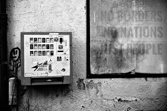 Black and white photo of a wall with a cigarette machine on the left and a frame on the top right.