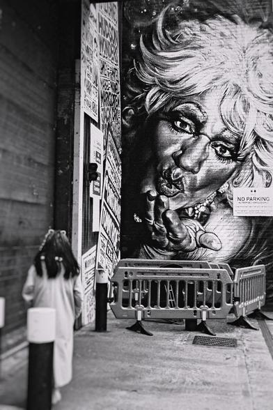 A black and white photograph of a young person walking past a large mural.