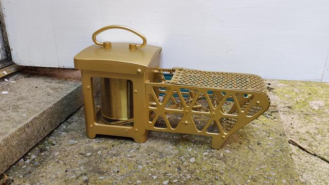 A strange looking mechanical object on a front step, propping open a white door. The contraption is golden in colour and consists of a chamber with a cylinder and a handle on top, and an elongated part with mesh and metal parts that might put you in mind of an old fashioned iron bridge.