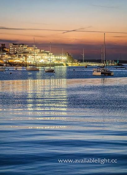 A very calm scene. Yachts moored on a flat sea, a town in the distance. It's dusk and the crescent moon is low in the sky, reflections from the artificially lit town are reflecting in the sea, rippling small waves add some interest .