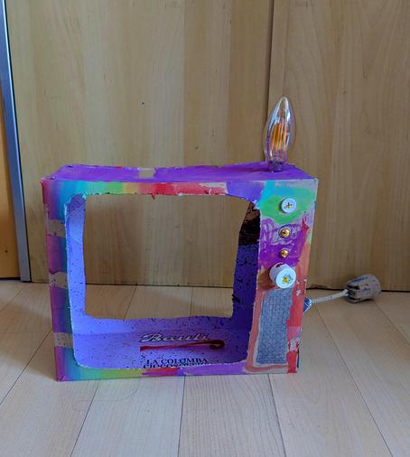 A brightly rainbow coloured cardboard cathode-ray TV, with buttons added to the cardboard, a bulb attached to the top as an antenna, and a wire hanging out the back.