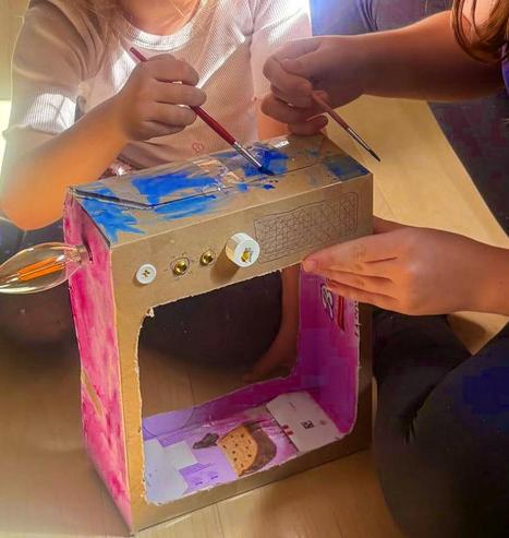 Kids painting a brightly rainbow coloured cardboard cathode-ray TV, with buttons added to the cardboard, a bulb attached to the top as an antenna, and a wire hanging out the back.