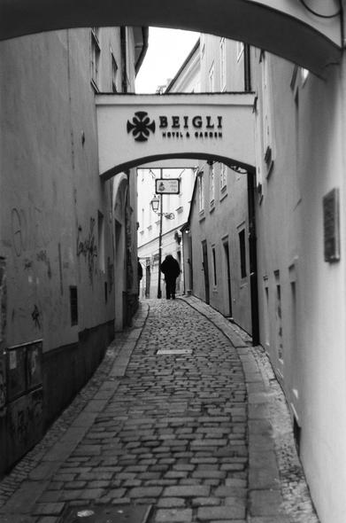 Black and white film photo of a dark figure walking down a cobble stone street. He has walked past an overhead piece advertising a hotel. Taken in Bratislava old town, Slovakia.