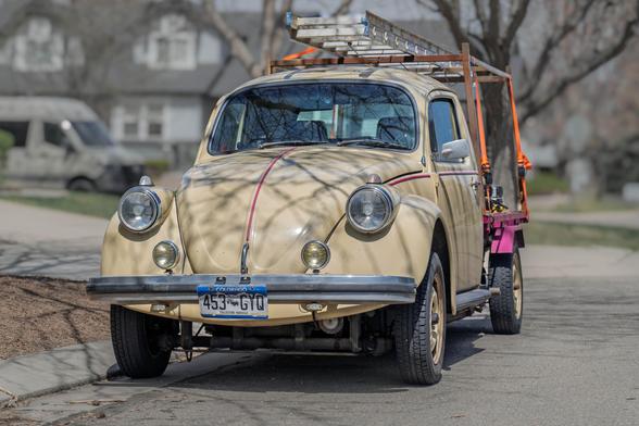 Front view of a modified vintage Volkswagen Beetle, painted beige with red pinstripes, converted into a pickup truck. A large ladder is strapped to a rack on the bed, and the vehicle is parked on a residential street.