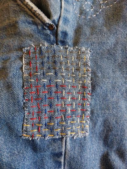 Flicken in Sashiko-Technik mit buntem Stickgarn auf einer Jeansnaht. Die Fäden bilden ein Webmuster.