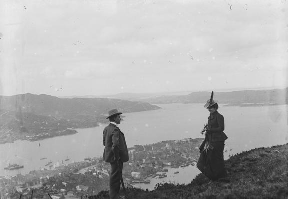 Black and white photo. In the foreground, a man and woman looking at each other. In the background, the view of Bergen seen from nearby mountain Fløyen, and the fjordscape towards Askøy Island.