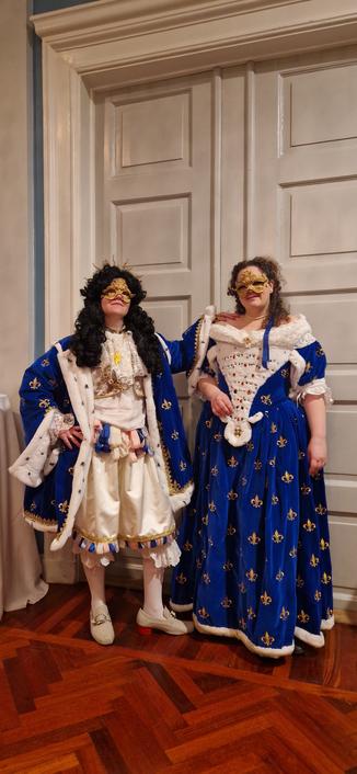 This photo shows two individuals in elaborate, matching blue and white costumes inspired by 17th–18th century French court fashion—specifically, the Baroque era associated with Louis XIV (the Sun King) and the court of Versailles.

The person on the left is dressed in a richly decorated blue robe with fleur-de-lis motifs, trimmed with white faux ermine, lace cuffs, and a wig—evoking royal attire or masquerade costume from the late 1600s. The gold mask and cravat complete the look.

The person on the right is wearing a gown in the same color and motif, also trimmed with white fur and adorned with jewels. The off-shoulder neckline, structured bodice, and full skirt align with the rococo-influenced court dresses of the early 1700s.