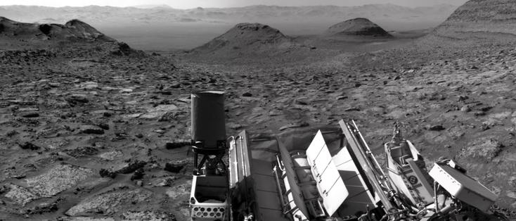A black and white image capturing a Martian landscape, with the rear section of the Curiosity rover in the foreground. The two buttes at center are Gould Mesa and Wilkerson Butte, with the wall of the rim of Gale Crater in the background, at 10 kilometers distance. Texoli Butte shows up on the right hand side.