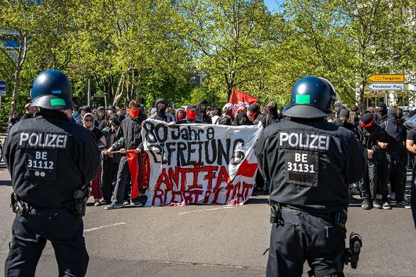 Das Foto von © Björn Obmann zeigt 2 behelmte Polizeibeamte die auf protestierende Menschen schauen. Diese halten ein Transparent mit dem Text: "80 Jahre Befreiung - Antifa bleibt ..."