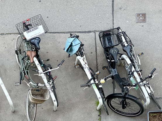 Bird's eye view of a residential street: several bicycles are parked at four bicycle parking spaces. A basket is attached to one of them, containing a folded newspaper with a large bold headline: “EDUCACIÓN !”.