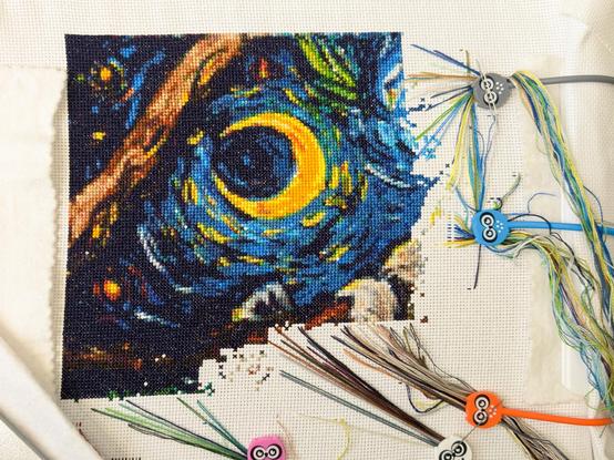 Full coverage cross stitch surrounded by a chaos of parked threads. The pattern is a swirly dark blue night sky with a big yellow crescent moon and some as-yet mysterious claws clinging to a tree branch.