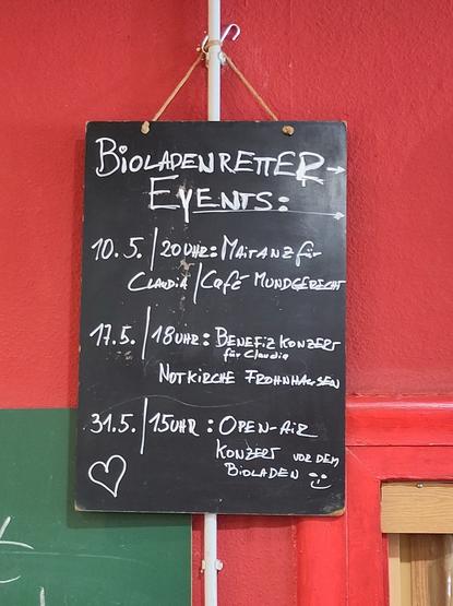 Schwarze Tafel mit Kreide beschrieben: 
Bioladen retter-Events: 
10.5. / 20 Uhr: Maitanz für Claudia / Cafe Mundgerecht 
17.5. / 18 Uhr: Benefizkonzert für Claudia / Notkirche Frohnhausen 
31.5. / 15 Uhr: Open-air-Konzert vor dem Bioladen