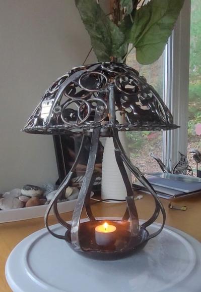 Sur la photo un champignon fait en rebut de métal qui sert de chandelier à la base et près du chapeau. 
Le chapeau est composé de toutes sortes d'objets (vis, bolt, ustensils, etc) qui, superposés et soudés ensembles, forment un tout disparate et plein de trous. 
Le corps du champignon est composé de barres de métal assez distanciées pour insérer une grosse chandelle bien que sur la photo il n'y a qu'un lampion qui soit allumé.