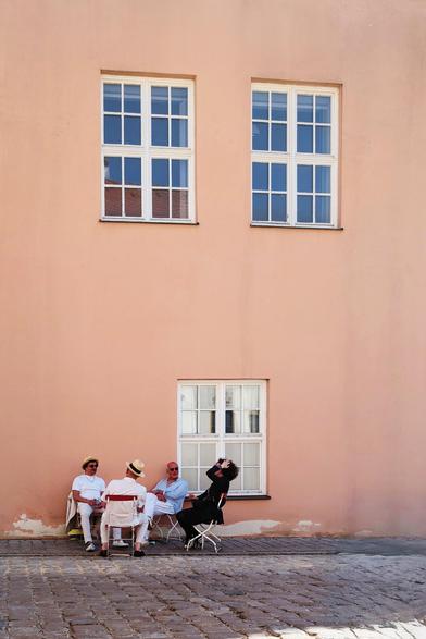 A group of people are sitting around a table in the bottom left of the photo in front of a wall with two windows in the top.
