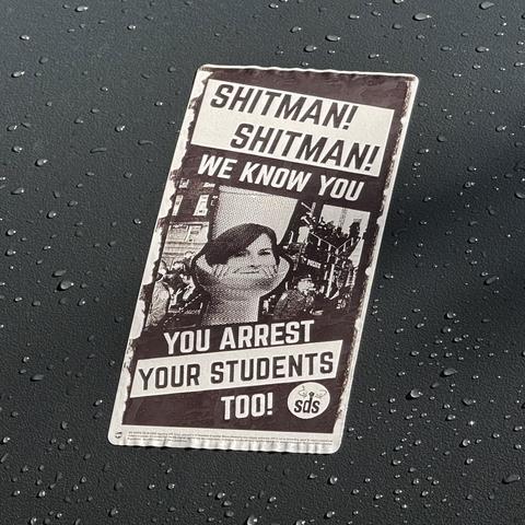 A sticker on the side of a traffic signal box on a lamp post reading “Shitman! Shitman! We know you — you arrest your students too!” showing a toilet with Claire Shipman’s (acting president of Columbia University) head in the toilet.