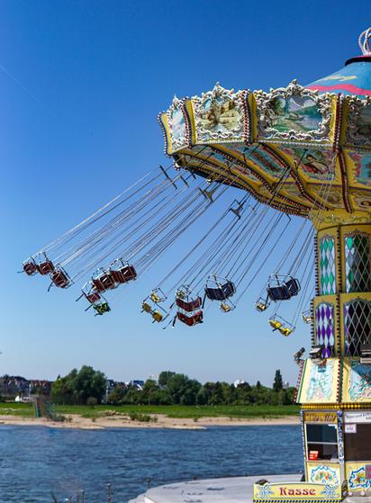 Eine farbenfrohe Kettenkarussell mit mehreren aufgehängten Sitzen vor einem klaren blauen Himmel. Das Kettenkarussell ist mit lebendigen Mustern und Bildern geschmückt, während im Hintergrund ein Fluss und eine grüne Landschaft zu sehen sind.

A colorful swing ride with multiple suspended seats, set against a clear blue sky. The ride is decorated with vibrant patterns and images, while a river and green landscape are visible in the background.