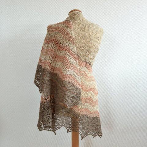 A knit shawl in cream, pink, and gray yarn with lace patterning, is shown from the back on a mannequin against a white background.